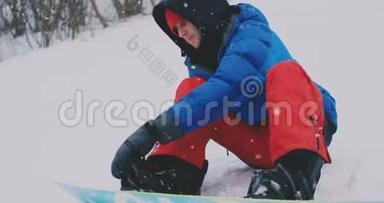 一个穿着红裤子的人坐在雪地上，在滑雪坡上系着滑雪<strong>板鞋</strong>和一件蓝色夹克